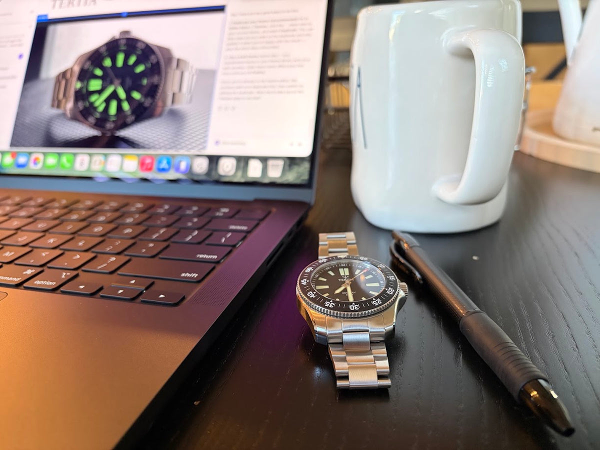A stainless steel analog watch with a black dial and bezel sits on a dark wooden desk next to a laptop, a white mug, and a pen.